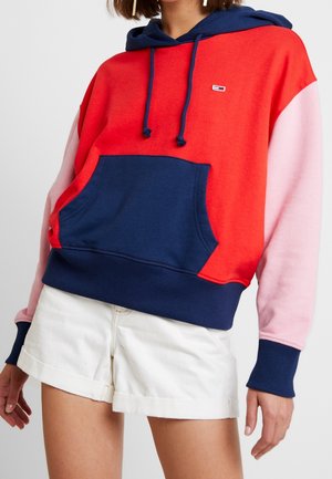 Vrouw draagt een color-block hoodie met een rode romp, een marineblauwe capuchon en zak, roze mouwen en witte korte broek, afgebeeld van hals tot midden dij.