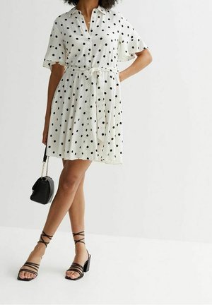 Vrouw die een witte korte mouwen jurk met polkadots draagt, zwarte sandalen met bandjes en een zwarte handtas met een kettingband vastheeft.