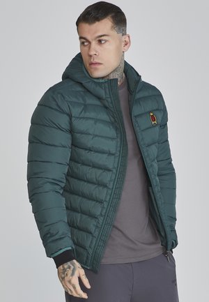 Chaqueta acolchada verde con capucha, que presenta un cierre de cremallera y puños acanalados. Logo bordado en el pecho. Lleva puesta una camiseta gris.