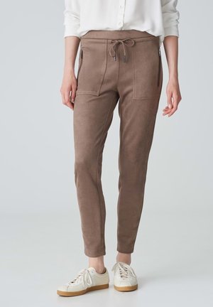 Pantalons de jogging marron en matériau doux et texturé, dotés de deux poches latérales zippées et d'une taille à cordon de serrage, associés à des baskets blanches.