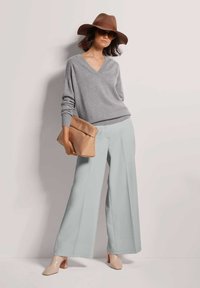 Frau mit einem breitkrempigen braunen Hut, Sonnenbrille, grauem V-Ausschnitt-Pullover, hellblauen weiten Hosen, beigen Stiefeletten mit Absatz und einer beige Clutch in der Hand.