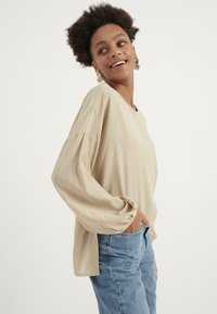 Hellbeige Langarmshirt mit lockerer Passform, runder Ausschnitt und Knopfdetails. Kombiniert mit blauen Jeans, die einen lässigen Look unterstreichen.