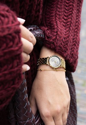 Montre en or à cadran rond et bracelet en métal, accompagnée d'un délicat bracelet en chaîne dorée, posée sur un pull bordeaux texturé.