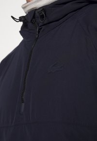 Mörkblå vindjacka med kvartsgång, prydd med en broderad logotyp på bröstet och justerbara snoddar på huvan.