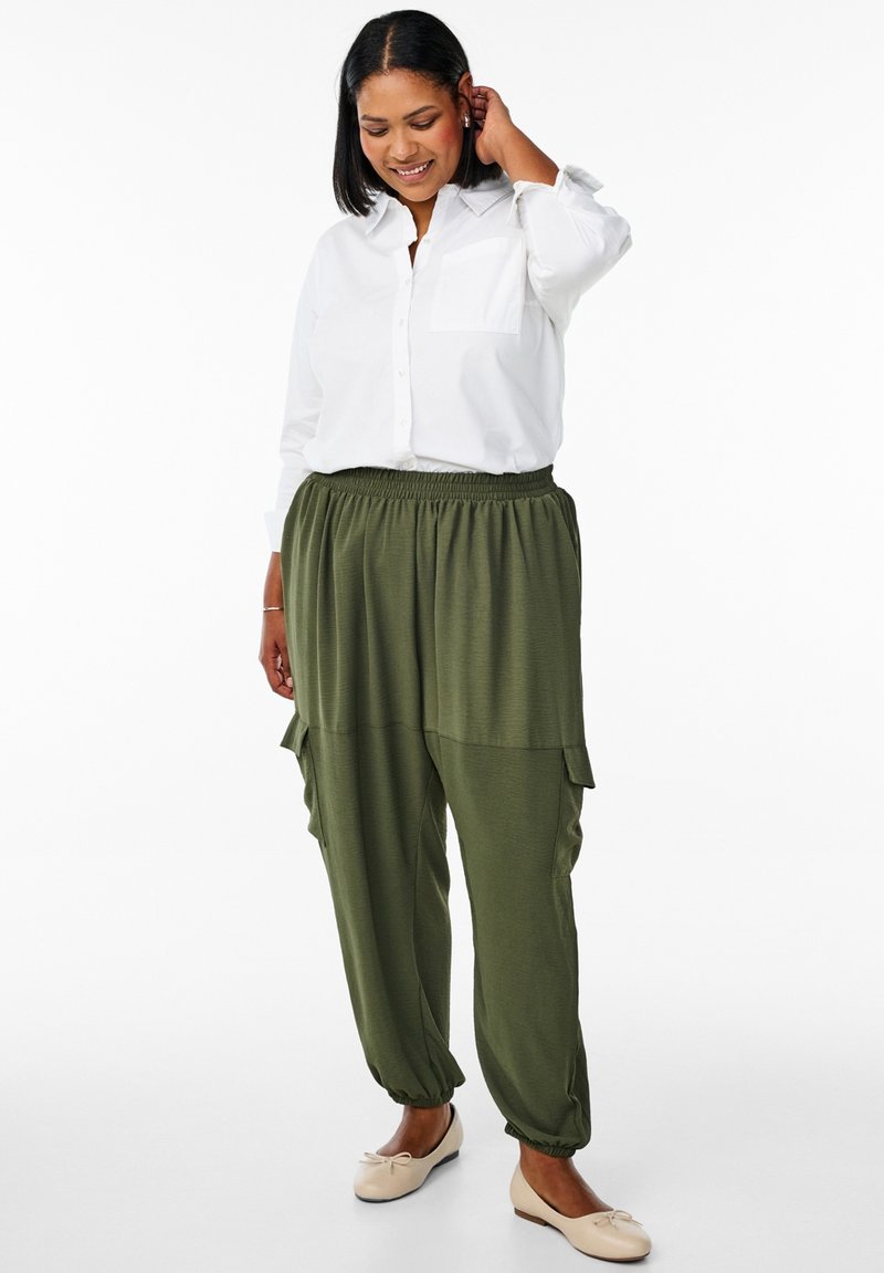 Femme souriante, la main touchant ses cheveux, portant une chemise blanche boutonnée, un pantalon ample vert olive avec poches et des ballerines beige, debout sur fond blanc.