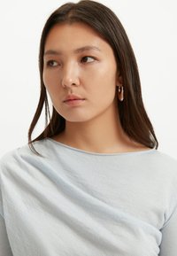 Hellblaues Oberteil aus weichem Stoff mit Bootsausschnitt und drapiertem Design. Das Modell trägt Creolen, hat glattes Haar und dezentes Make-up.