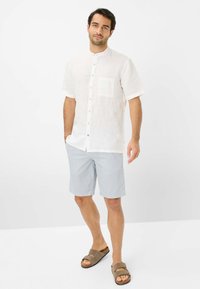 Camicia bianca in lino a maniche corte con chiusura a bottoni, taschino sul petto, abbinata a pantaloni corti a righe blu chiaro e sandali marroni.