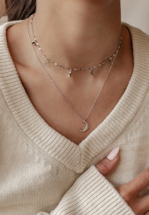 Silberne geschichtete Halsketten mit Mondanhängern in verschiedenen Größen und Designs, getragen über dem Halsausschnitt eines leichten gestrickten Pullovers.