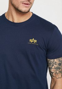 Marineblå bomull t-skjorte med en liten gul logo på venstre bryst, rund hals og korte ermer. Tettsittende design med glatt tekstur.