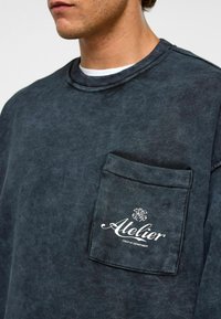 Donkerblauwe sweatshirt met een zak en witte "Atelier" tekst. Beschikt over een ronde hals, zachte textuur en subtiel verouderd ontwerp.