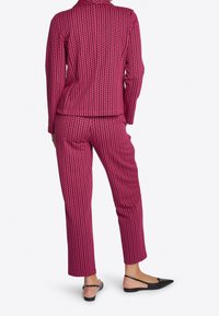 Roze pak met patroon, bestaande uit een getailleerd colbert en taps toelopende broek. De stof heeft een gestructureerde weving. Gekleed met zwarte platte schoenen, gezien vanaf de achterkant.