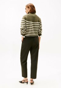 Woman standing sideways wearing olive green and white striped turtleneck sweater, dark pinstriped pants, and black kitten heels.