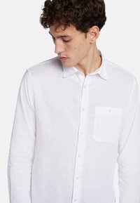 Chemise blanche en lin à boutons avec une poche, dotée d'un col pointu et de boutons visibles, mettant en valeur un tissu texturé et léger.