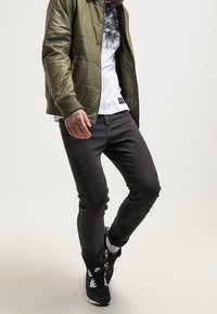 Olivgrüne Steppjacke, weißes Grafik-T-Shirt, ausgefranste schwarze Jeans und schwarze Sneaker. Texturierte Materialien mit lässigem Streetwear-Design.