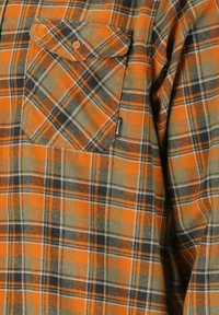 Flanellikosketi oranssissa, mustissa ja harmaissa ruuduissa. Siinä on napitettava rinta-tasku ja pehmeä, teksturoitu kangas.