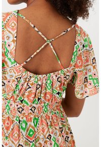 Mujer vestida con un vestido de estampado colorido, con tirantes delgados entrecruzados y un detalle de espalda elástica, que muestra su parte superior de la espalda y su cabello rizado.