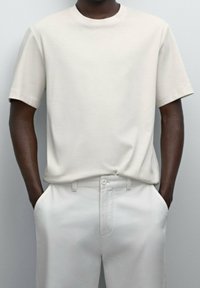 Mörkhyad person som bär en enkel ljusgrå t-shirt med kort ärm, instoppad i matchande högmidjade byxor och med händerna i fickorna.