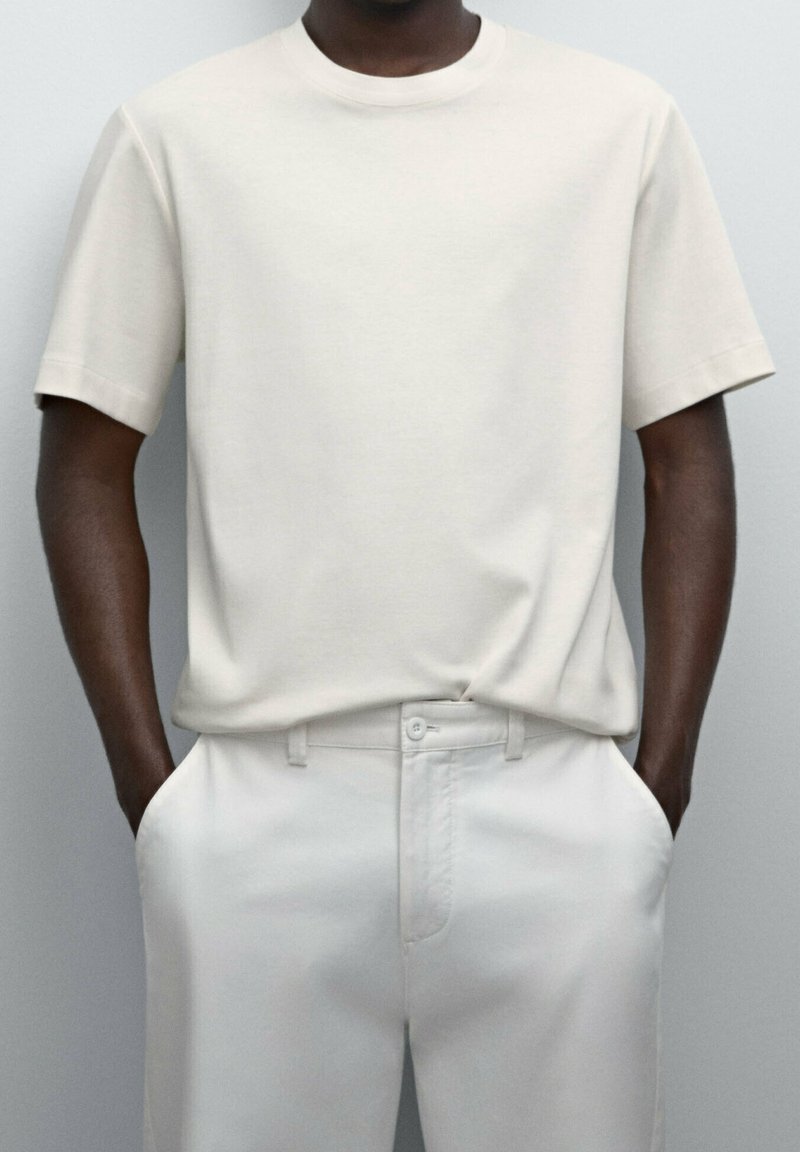 Mörkhyad person som bär en enkel ljusgrå t-shirt med kort ärm, instoppad i matchande högmidjade byxor och med händerna i fickorna.