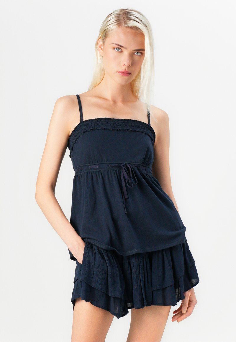 Top sin mangas azul marino con cuerpo fruncido y tirantes ajustables, dobladillo de volantes en capas y detalle de lazo. Textura de tela suave.