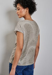 Top de manga corta de un ligero tono beige con un sutil patrón texturizado. Corte relajado, escote curvado y detalle de pequeña abertura lateral. Combinado con jeans azules.
