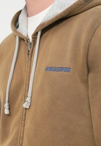 Καφέ φούτερ με φερμουάρ, με γκρι φόδρα κουκούλας, διαθέτει γκρι κορδόνια και κεντημένο λογότυπο "Hollister" σε μπλε χρώμα.