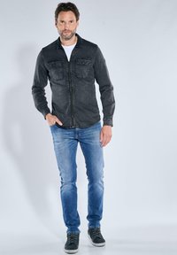 Mann in schwarzer Zip-Jacke, weißem Hemd, blauen Jeans und schwarzen Sneakers, der mit einer Hand in der Tasche vor einem einfachen hellen Hintergrund steht.