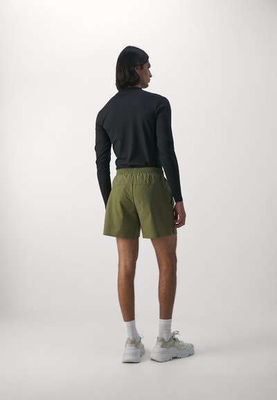 Haut noir à manches longues, shorts verts avec une taille élastique et baskets légères. Les shorts ont une texture lisse et une coupe décontractée.