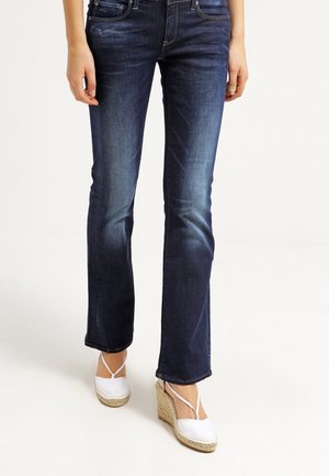 Femme portant un jean bootcut bleu foncé et des espadrilles compensées blanches, debout sur un fond blanc.