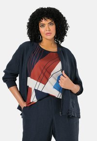 Giacca blu navy con texture e polsini a coste, abbinata a un top con motivo geometrico multicolore che presenta accenti rossi, blu e beige.