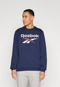 Tamno plavi dukserica s okruglim izrezom, dugim rukavima i bijelim Reebok logotipom s crvenim naglascima. Izrađena od mekog materijala, s rebrastim manžetama.
