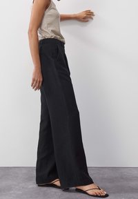 Mustat leveät housut, joissa on korkea vyötärö, näyttävät sileältä kankaalta ja puhtaalta siluetilta. Yhdistetty beigeen hihattomaan puseroon.