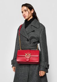 Červená kožená kabelka cez telo so zlatým logom na sponke, obdĺžnikový dizajn a odnímateľný reťazový popruh. Hladký povrch a lesklý finiš.