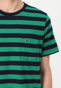 Vihreä ja laivastonsininen raidallinen T-paita, lyhyet hihat, pyöreä kaula-aukko, jossa on rintatasku pienen logovetin kanssa vasemmalla puolella. Puuvillamateriaalia.