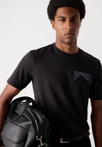 Black short-sleeve t-shirt featuring a logo on the chest. The model holds a black leather bag with visible stitching and silver zipper.