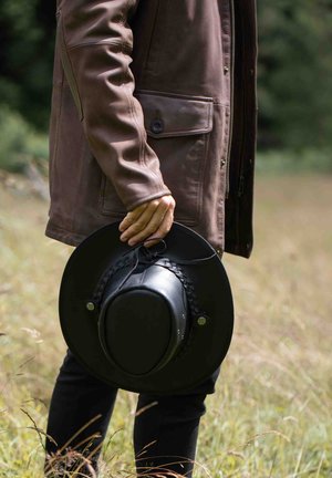 Black felt hat with braided trim held by a person wearing a brown leather jacket, standing in a grassy field under natural light.
