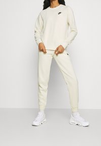 Cream-colored Nike sweatshirt and matching joggers. Soft fabric, ribbed cuffs, and hem with a small black logo on the chest and pants. White sneakers.