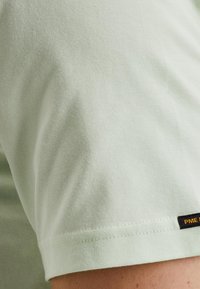 Lys grøn bomulds t-shirt stof med en glat tekstur. Har et sort mærke med hvid tekst på kanten. Nærbillede af ærmet.