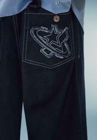 Jeans din denim închis, cu un design distinctiv brodat sub formă de stea și cerc pe buzunarul din spate, având un detaliu din cupru pe talie.
