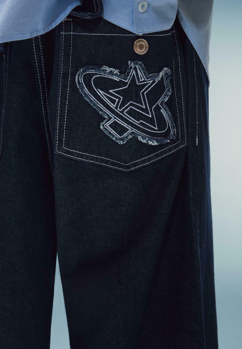 Jeans din denim închis, cu un design distinctiv brodat sub formă de stea și cerc pe buzunarul din spate, având un detaliu din cupru pe talie.