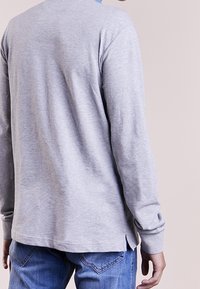 Maglietta a maniche lunghe grigia chiaro in tessuto morbido, con aperture laterali all'orlo e scollatura rotonda. Jeans di denim visibili in fondo.