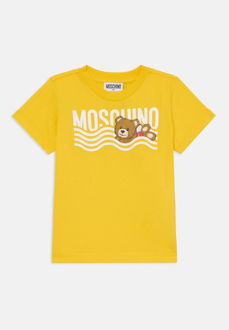 Žluté bavlněné tričko s grafikou medvěda a nápisem "MOSCHINO" v bílé barvě, s vlnitými liniemi v pozadí. Krátké rukávy.