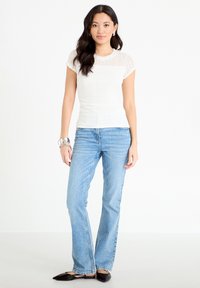 Joven mujer de pie con una blusa blanca de encaje de manga corta, vaqueros azul claro, pulsera plateada y zapatos planos negros puntiagudos.