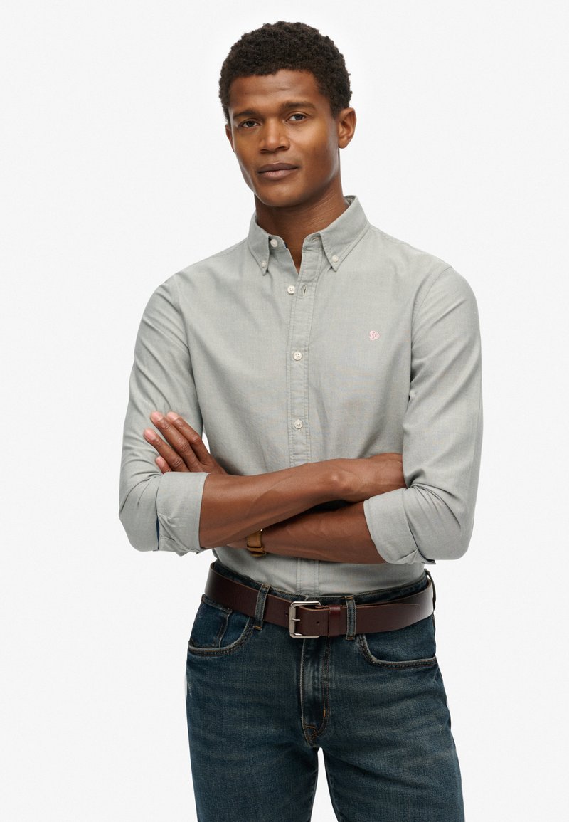 Camisa de botón gris claro con cuello, mangas enrolladas y un sutil logo rosa, combinada con jeans de mezclilla oscuros y un cinturón marrón.