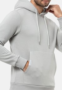 Hellgrauer Hoodie mit einer Fronttasche, Kordelzugkapuze und gerippten Bündchen. Weicher Stoff mit glatter Textur und minimalen Designdetails.