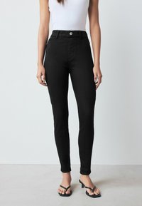 Aukštos juosmens juodos džinsai slim fit stiliaus, pagaminti iš lygaus džinsinio audinio. Yra priekiniu mygtuku ir kišenėmis. derinami su juodais šlepetėmis.