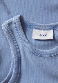 Camisola de alças azul claro com textura canelada, apresentando uma gola arredondada e um rótulo com "JJXX" bordado em marinho. A textura é suave e ligeiramente elástica.