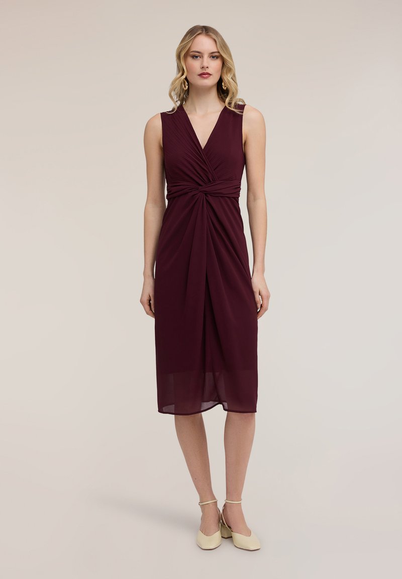 Kvinde iført ærmeløs, knælængde bordeaux kjole med snoet frontdetalje og beige ankelrem hæle, stående mod en ensfarvet baggrund.
