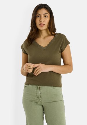 Mujer con cabello largo y oscuro, vestida con una blusa verde oliva con escote en V y detalles de encaje, y pantalones verde claro, de pie con las manos juntas.