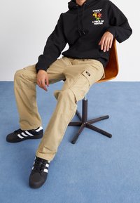 Černá mikina s kapucí s grafickou potiskem na hrudi, béžové cargo kalhoty s kapsami a černé tenisky s bílými pruhy, sedící na dřevěné židli.