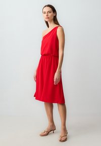 Vrouw in een knielange, rode, one-shoulder jurk met gouden gespsdetails, beige, hoge sandalen en armband, staand tegen een effen lichte achtergrond.